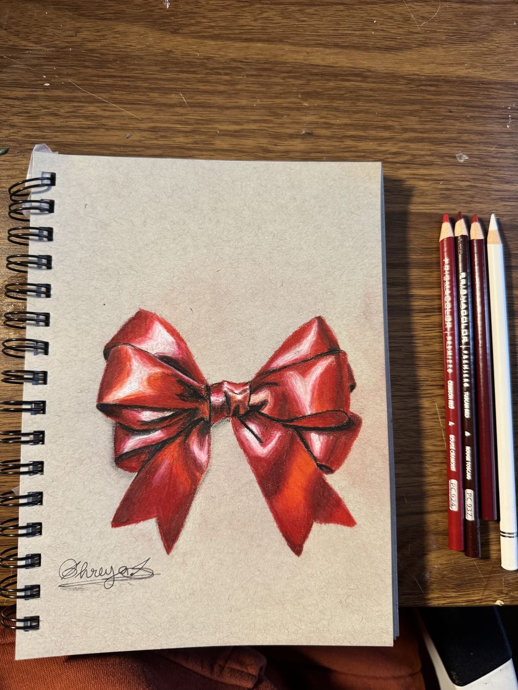 Coloured-pencil drawing of a red satin bow in a sketchbook, pencils alongside.