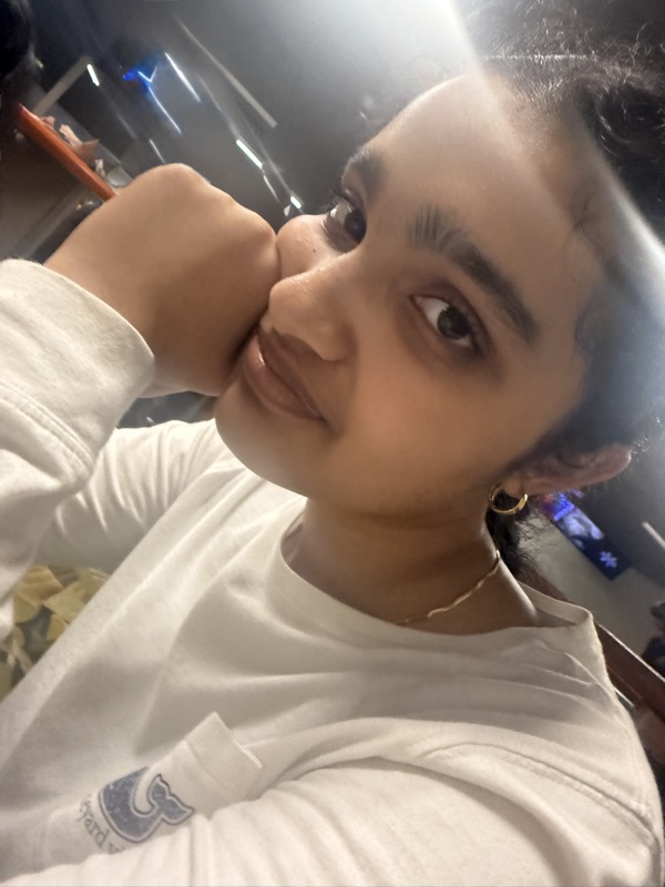Shreya resting her chin on her hand in a white sweatshirt, close up, with coloured lights blurred behind her.
