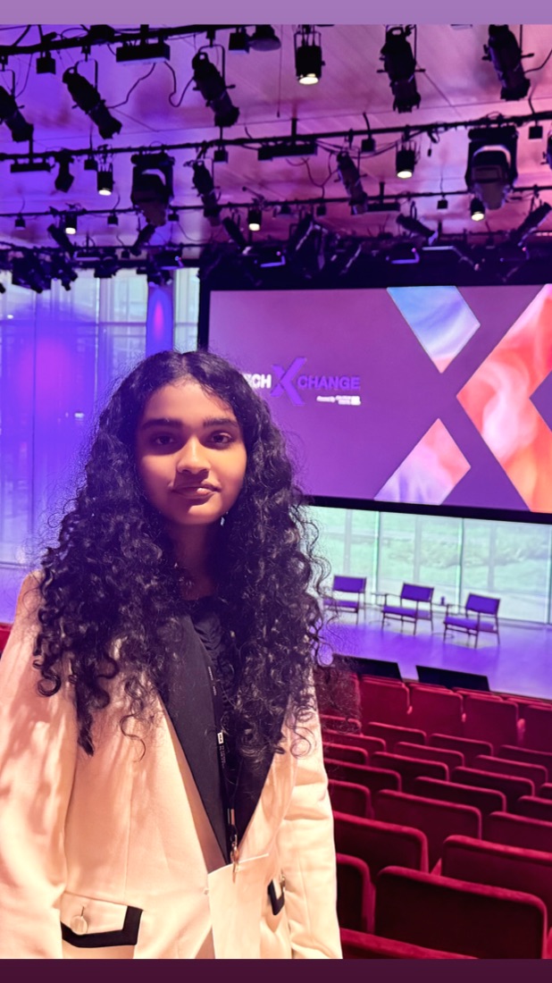 Shreya standing in a theatre auditorium in a cream blazer, with a TechXchange stage screen lit purple behind her.