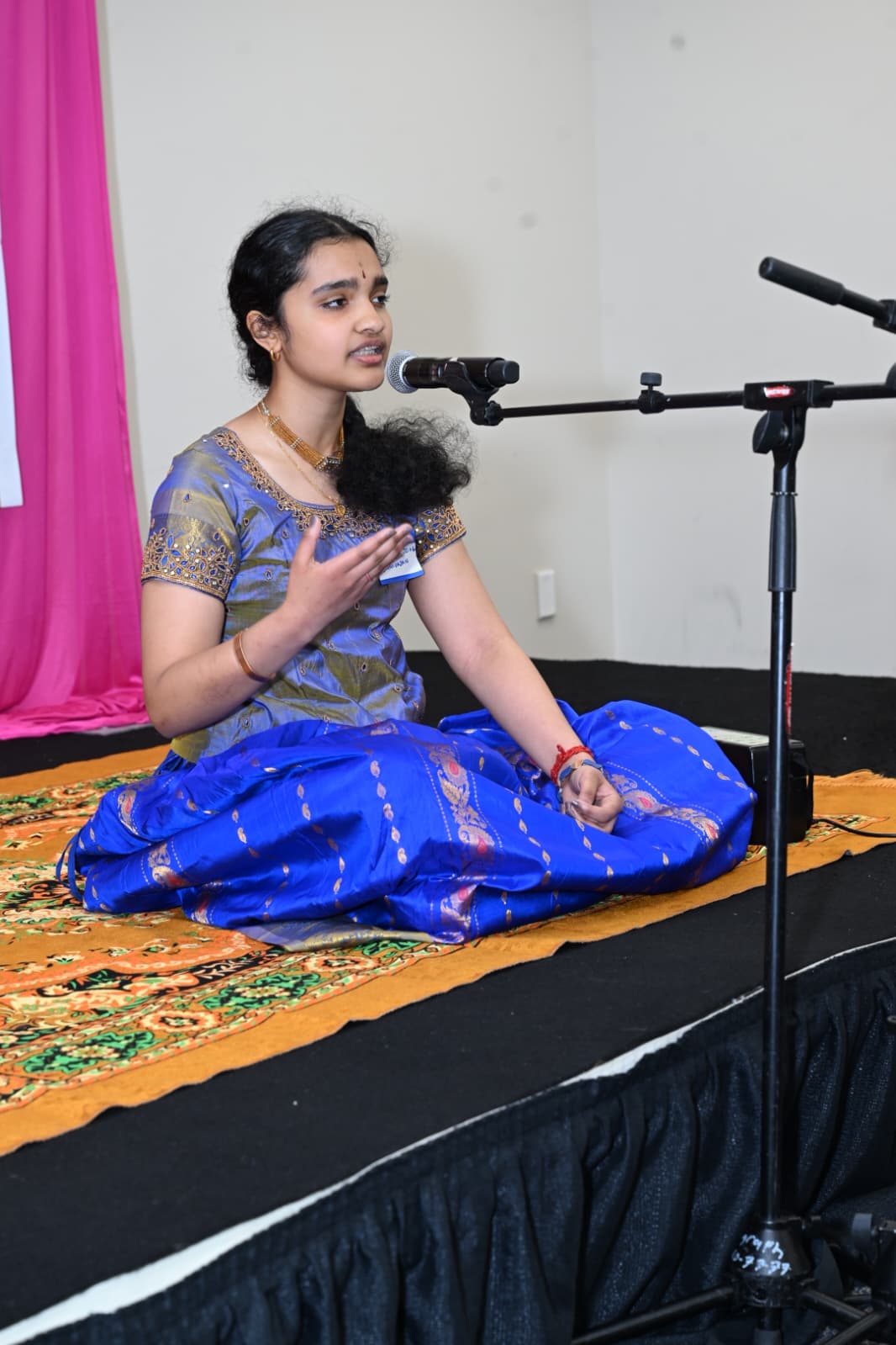 Shreya seated cross-legged on stage in a blue and gold silk sari, singing into a microphone during a classical vocal recital.